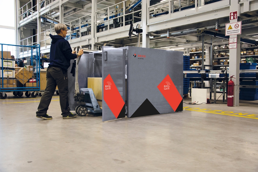 Eine dunkel gekleidete Mitarbeiterin schiebt die Palette aus dem RFID Gate in einem großen Industrielager mit Regalen, Förderbändern und verschiedenen Geräten im Hintergrund. Das RFID-Gate ist grau mit roten und schwarzen Akzenten.